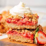Two stacked strawberry oatmeal bars topped with whipped cream rest on a wooden board, accompanied by a fresh strawberry slice nearby.