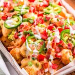 A tray of loaded tater tot nachos piled high with melted cheese, sliced jalapeños, diced tomatoes, chopped bacon, and green onions.