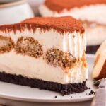 A slice of layered tiramisu cheesecake sits invitingly on a plate with a fork. The cake boasts a cocoa-dusted top with creamy, coffee-soaked layers that promise indulgence with every bite.