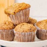 A plate of five zucchini muffins, one perched atop the others, set against a kitchen background with a checkered cloth partially visible.
