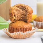 A stack of banana muffins graces the table, one enticingly sliced open to reveal its moist interior. A green stand artfully complements the display, while a glass of milk waits invitingly in the background.