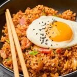 A bowl of spicy kimchi fried rice topped with a sunny-side-up egg and sprinkled with black sesame seeds, served with chopsticks.