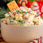 A festive bowl of Christmas tree cake dip topped with sprinkles and a graham cracker, surrounded by pretzels and holiday decorations.