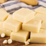 A pile of square pieces of vanilla fudge is stacked on a wooden surface, surrounded by scattered white chocolate chips. A blue and white striped cloth is partially visible in the background.