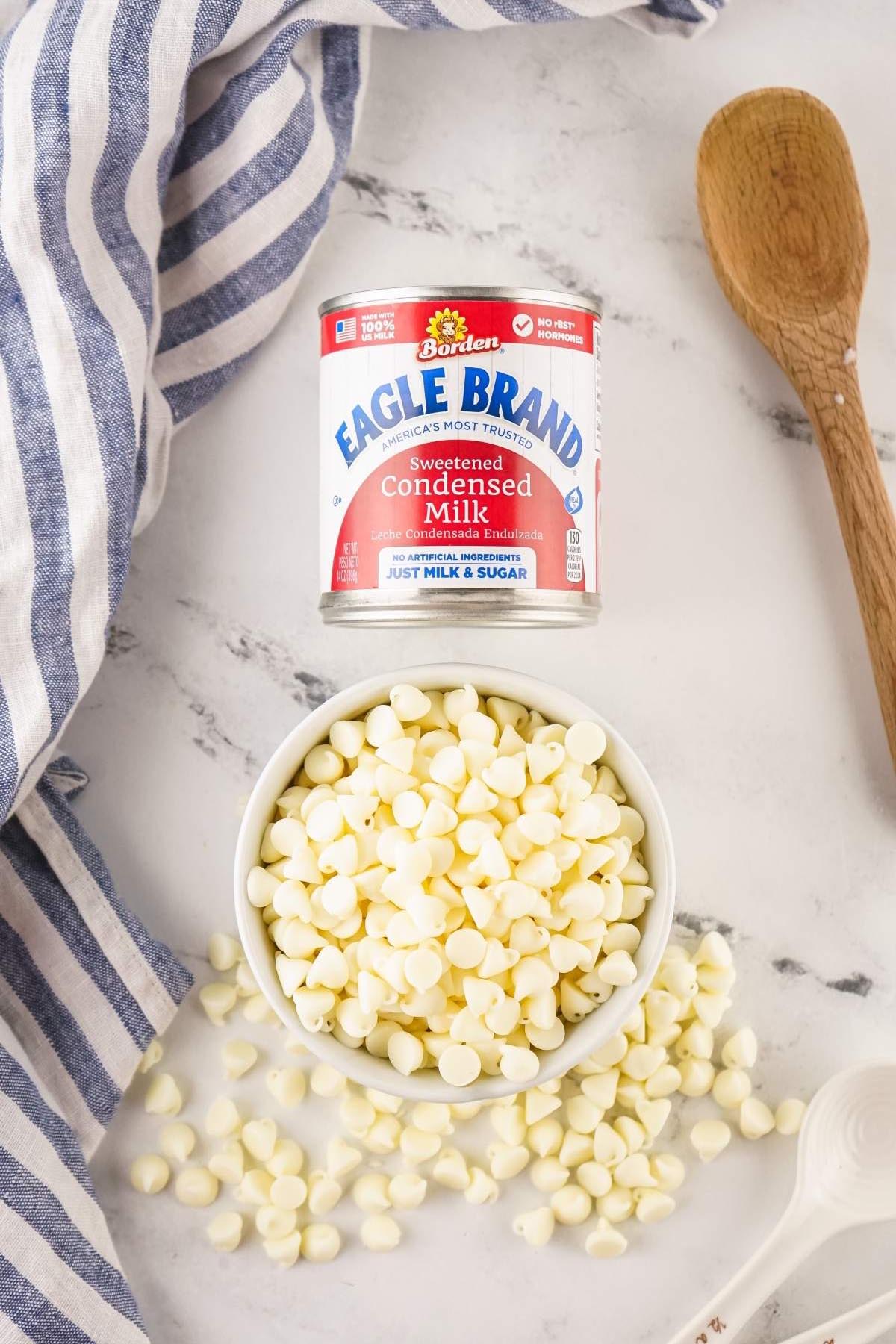 Bowl of white chocolate chips with spilled chips, a can of sweetened condensed milk&mdash;perfect for making easy 2-ingredient vanilla fudge&mdash;plus a striped towel and wooden spoon.
