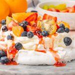 An air fryer pavlova topped with blueberries, strawberries, kiwi, and orange slices sits elegantly on a marble surface. Bowls of additional fruit are in the background, ready to embellish this delightful creation even further.