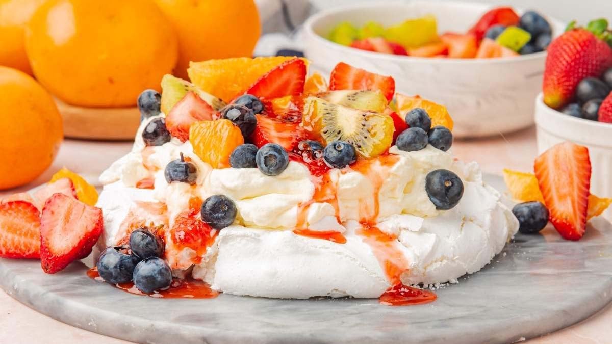 A pavlova topped with whipped cream, strawberries, blueberries, kiwi, and orange slices on a marble surface, surrounded by whole strawberries and oranges.