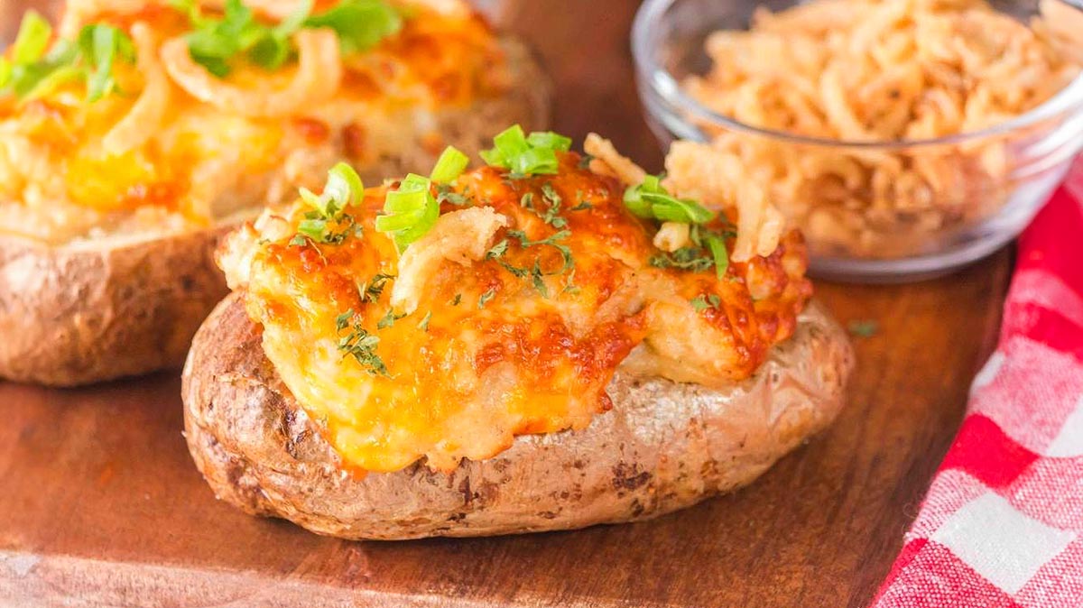 Loaded baked potatoes topped with melted cheese, green onions, and crispy fried onions, served on a wooden surface with a small bowl of more crispy onions in the background.
