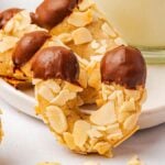 Crescent-shaped chocolate-dipped almond horn cookies are topped with sliced almonds and elegantly arranged on a white tray, accompanied by a refreshing glass of milk.