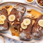 Four slices of banana bread French toast topped with banana slices, pecans, syrup, and powdered sugar on an oval plate. Surrounding items include extra syrup, crunchy nuts, and a refreshing glass of milk.