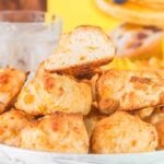 A plate of Bisquick cheddar biscuits stacked with a halved cheddar biscuit on top. Two glasses of ice water and a colorful cereal box are in the background.