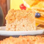 A slice of a classic Bisquick coffee cake, sits on a plate with pancakes and berries artfully arranged in the background. A refreshing glass of milk is nearby, completing this delightful breakfast tableau.
