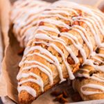 Two cinnamon scones with white icing drizzle and small brown pieces are placed on a parchment-lined tray.