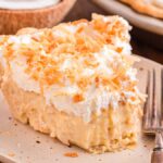 A slice of delectable coconut cream pie, adorned with a swirl of whipped cream and sprinkled with toasted coconut flakes, sits invitingly on a plate beside a fork.