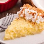 A slice of coconut custard pie sits elegantly on a plate, topped with whipped cream and sprinkled with toasted coconut flakes, alongside a ready fork.