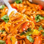 A fork lifts a portion of Thai drunken noodles stir-fry with vegetables and peanuts from a pan.