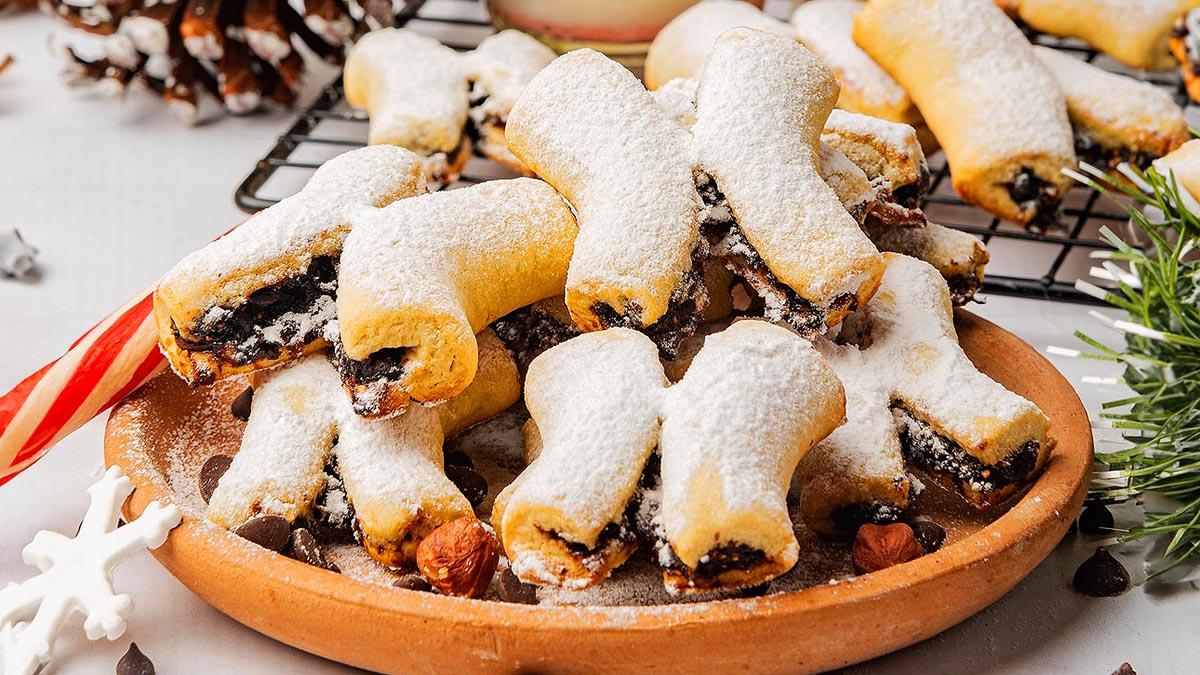 A plate of crescent-shaped pastries dusted with powdered sugar, filled with a dark jam or fruit mixture, placed on a wooden surface.