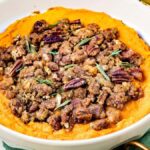 Bowl of maple garlic sweet potato casserole topped with pecans and herbs, resting on a green cloth.