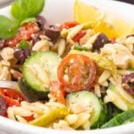A white bowl brimming with a Mediterranean tuna orzo salad features cherry tomatoes, cucumber slices, olives, and basil, all crowned with a lemon wedge. A fork rests beside the vibrant mix.
