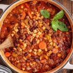 A pot of hearty beef and vegetable pasta e fagioli soup (Olive Garden copycat) with a wooden ladle, garnished with basil. A bowl of red pepper flakes and rustic bread sit nearby on a wooden table, creating the perfect setting for a comforting meal.