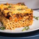 A slice of pastitsio, rich with cheese, ground beef, and vegetables, rests on a white plate garnished with chopped herbs. A fork is placed beside the hearty slice.