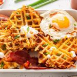 A plate of savory cheesy potato waffles topped with bacon, a sunny-side-up egg, diced green onions, and sour cream.