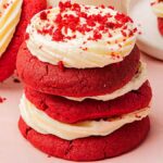 A stack of red velvet sugar cookies with cream cheese frosting and red crumbs on top, delicately arranged on a light pink surface.
