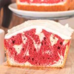A delectable slice of red velvet marble loaf cake with swirls of rich color, topped with velvety white frosting, rests invitingly on a wooden board. In the background, a steaming coffee pot and cup await to complete the perfect indulgent pairing.