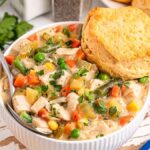 A bowl of slow cooker chicken pot pie soup brimming with vegetables, garnished with parsley, and topped with a biscuit. A spoon rests inside the bowl. Fresh parsley and a checkered cloth are visible nearby, adding to the cozy setting.