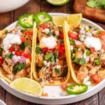 Three chicken tacos filled with slow cooker shredded Mexican chicken and topped with tomato, onion, cilantro, and sour cream on a plate. Lime wedges and bowls of garnishes surround the dish.