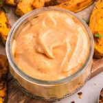 A glass bowl of spicy mayo sits on a wooden board alongside seasoned sweet potato wedges and a small bowl of chili flakes.
