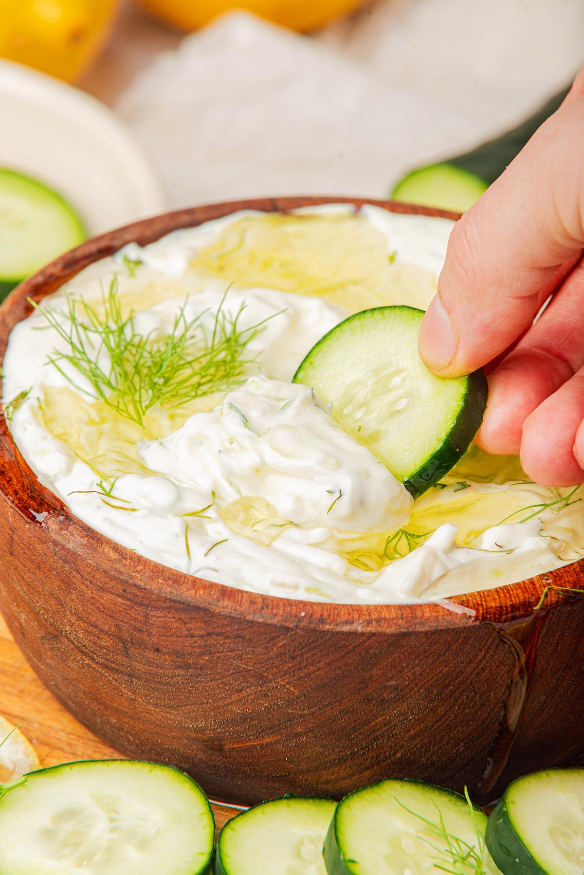 A hand dips a cucumber slice into a wooden bowl of Greek tzatziki dip, garnished with dill and drizzled with olive oil. Sliced cucumbers are scattered around, completing the fresh presentation of this classic tzatziki recipe.