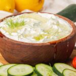 A wooden bowl of homemade tzatziki is surrounded by fresh cucumber slices, tomatoes, dill, and lemon.