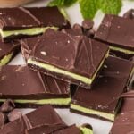 A pile of homemade Andes mints with green mint layers, surrounded by chocolate chips and fresh mint leaves.