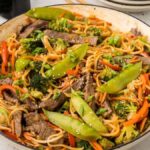 A pan of beef lo mein featuring stir-fried noodles with tender beef, snow peas, broccoli, and carrots, all garnished with sesame seeds and chopped green onions.