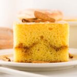 A slice of yellow honey bun cake with a cinnamon swirl, topped with cinnamon sticks, sits on a white plate.