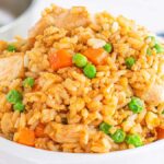 A bowl of chicken fried rice, packed with savory pieces of chicken, vibrant peas, and crisp carrots, accompanied by a wooden spoon.