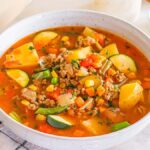 A bowl of hearty ground turkey vegetable soup with zucchini, potatoes, peas, carrots, corn, and savory herbs. A striped cloth and a small dish of fresh herbs are placed beside the bowl.