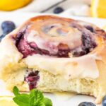 A glazed lemon blueberry sweet roll with berry filling sits elegantly on a white plate, garnished with fresh lemon slices, juicy blueberries, and sprigs of mint.