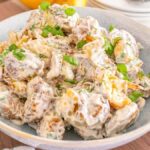 A bowl of smashed potato salad topped with chopped green onions rests on a wooden table.