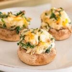 Three spinach artichoke stuffed mushrooms on a white plate, topped with a savory blend of cheese and herbs.