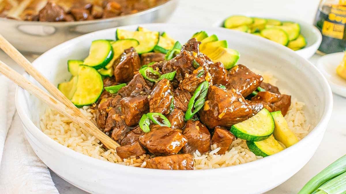 A bowl of rice topped with cubed steak bites in a brown sauce, garnished with sliced green onions and zucchini.