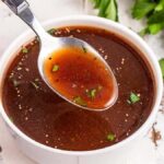 A spoonful of clear brown beef au jus with herbs hovers above a white bowl on a white surface, next to a bunch of parsley.