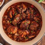 Pot of Jamaican brown stew chicken with vegetables on a wooden table, surrounded by a bowl of rice and beans, a wooden spoon, and fresh vegetables.
