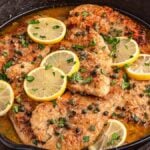 Pan-seared chicken piccata cutlets in a lemon butter sauce, garnished with lemon slices, capers, and fresh parsley, served in a black skillet.