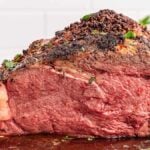 Close-up of a sliced, herb-crusted prime rib seasoned to perfection, displayed on a wooden board and garnished with fresh rosemary and parsley in the background.
