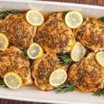 Lemon rosemary chicken thighs are roasted to perfection, topped with fresh herbs and lemon slices in a white baking dish, and beautifully garnished with delicate rosemary sprigs.