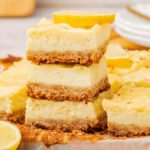 Lemon cheesecake bars stacked on a rustic wooden surface, beautifully garnished with fresh lemon slices, while plates and cutlery subtly frame the background.