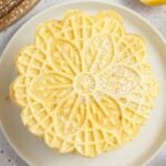 A stack of lace-patterned lemon pizzelle cookies dusted with powdered sugar sits on a plate, surrounded by fresh lemon slices and more cookies on a textured surface.