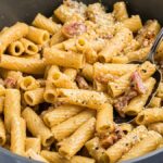 A bowl of rigatoni pasta alla gricia mixed with a creamy sauce, garnished with grated cheese and black pepper, has a spoon resting invitingly in the dish.